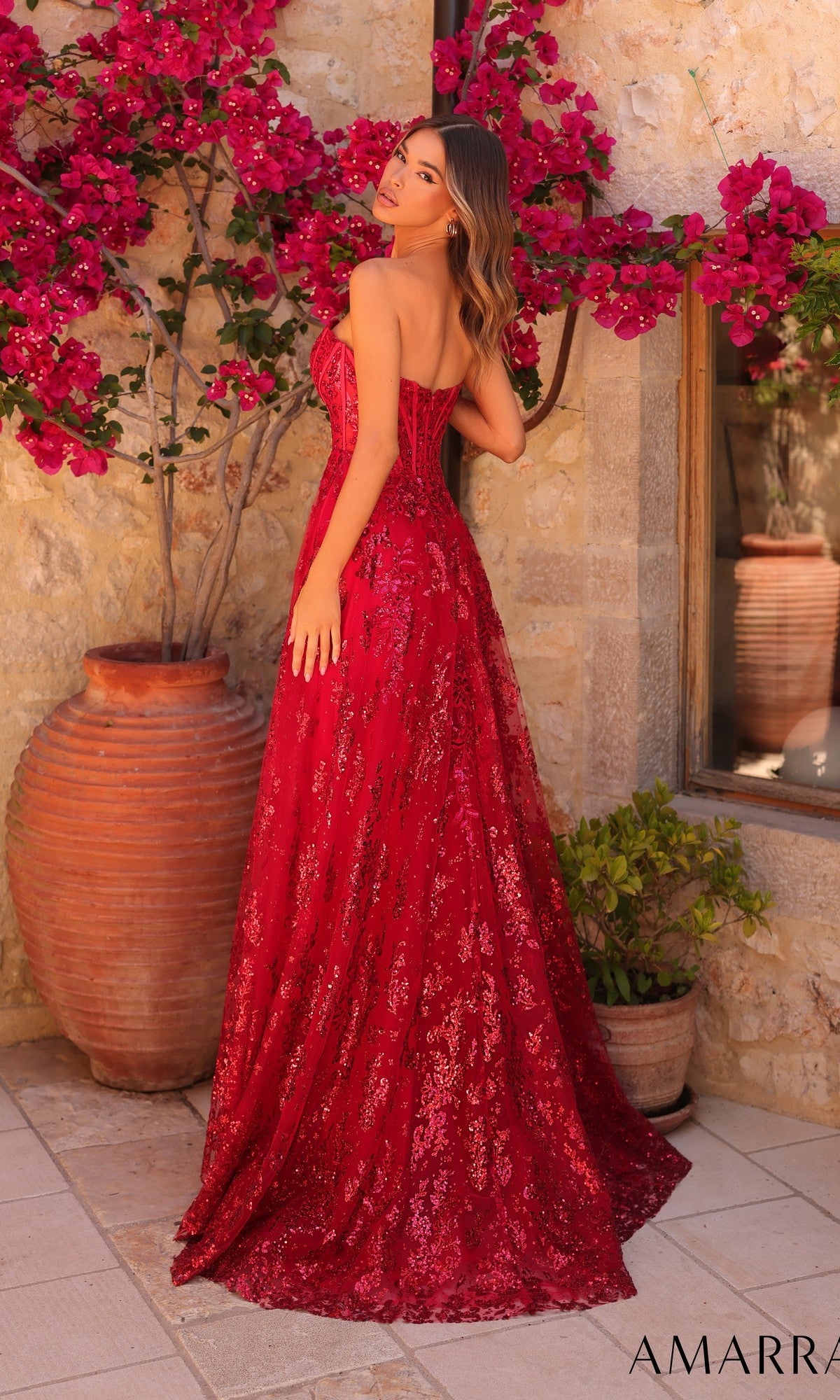 A woman wearing the Amarra 89074 long prom dress stands outdoors by a stone wall, with a large clay pot filled with pink flowers in the background.